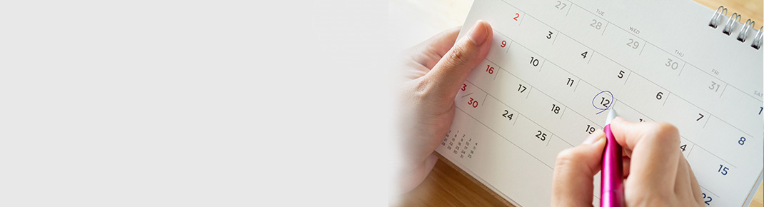 Vrouw in bril met donkerblauw shirt leunt op een wit bed met een glimlach, peinzend kijkend. / Close-up van een kalender met een vrouwenhand die een dag markeert met een pen.